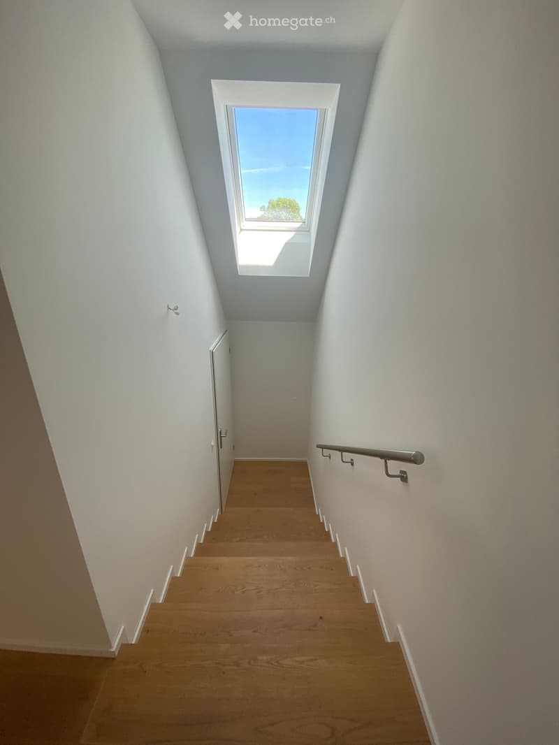 A narrow staircase with wooden steps and white walls, featuring a metal handrail on one side and a skylight at the top providing natural light.