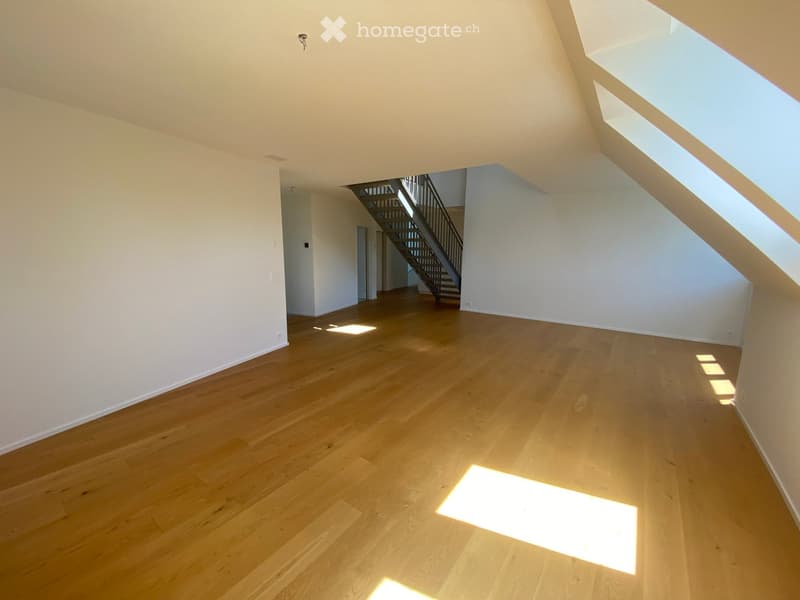 Empty room with wooden flooring and white walls, featuring a staircase leading to another level and large windows allowing natural light to enter.