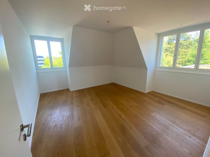 Empty room with wooden flooring, white walls, and large windows offering a view of greenery outside.