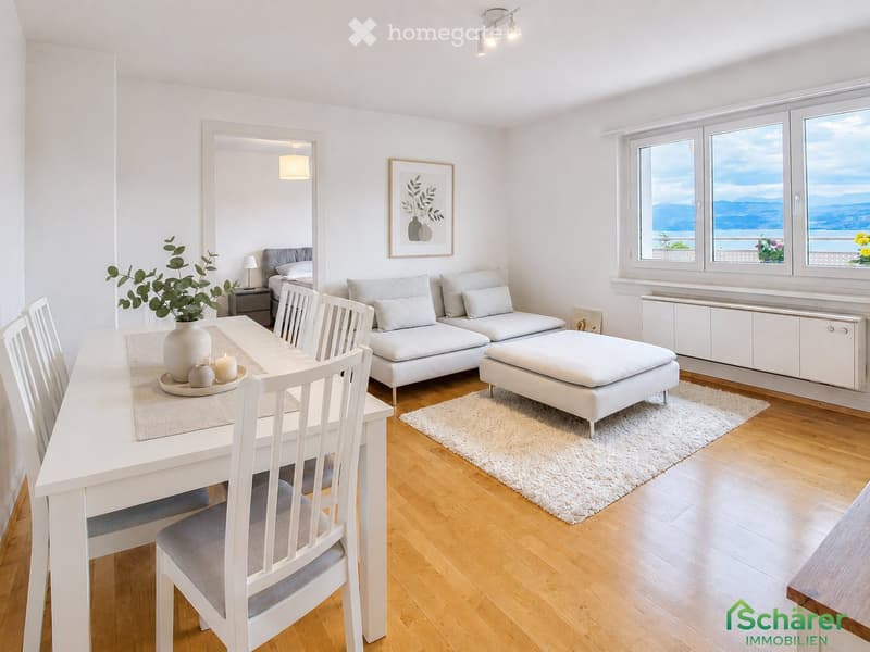 Bright, modern living and dining area with white walls, wooden flooring, large windows offering a scenic view, a white sofa with cushions, an ottoman, a dining table with chairs, a vase with greenery, and a mirror.