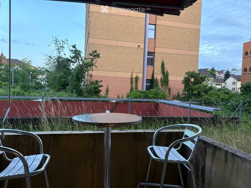 Balcony with a round table and two metal chairs, overlooking a fenced area with tall grass and trees, with a modern building in the background.