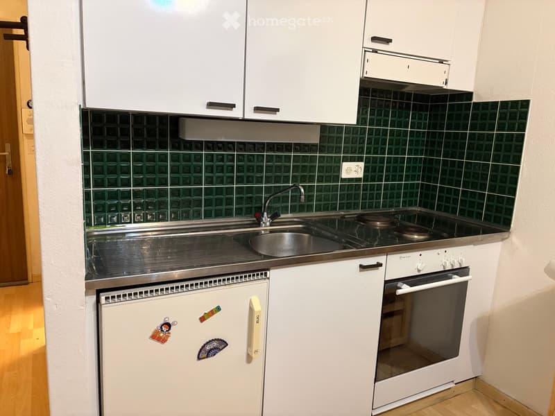 Compact kitchen with white cabinets, stainless steel sink, oven, and refrigerator. Green tiled backsplash and wooden flooring.