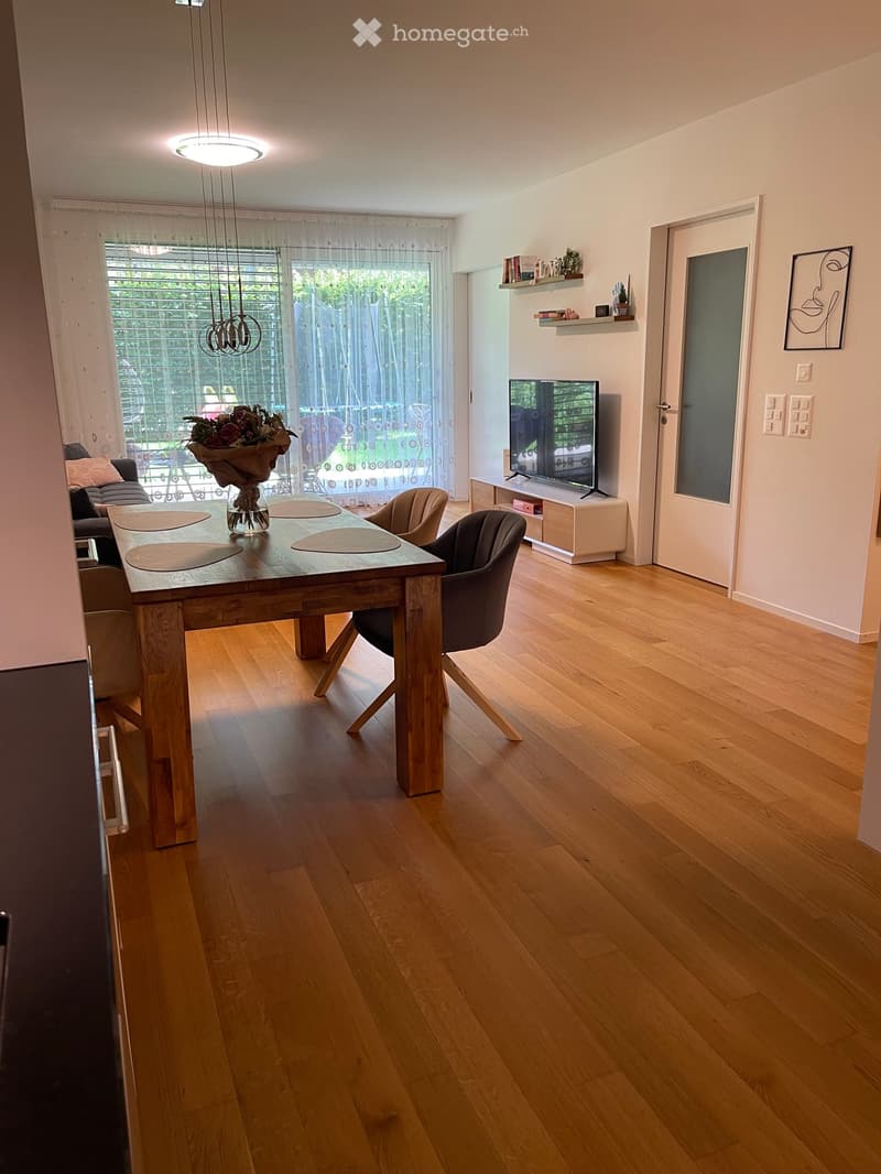 Bright living room with wooden flooring, large window with sheer curtains, a wooden dining table with chairs, a sofa, a television on a stand, and decorative shelves.