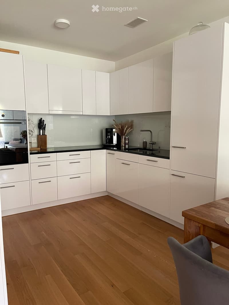 Modern kitchen with white cabinets, wooden flooring, stainless steel appliances, and a small dining area with a table and chair.