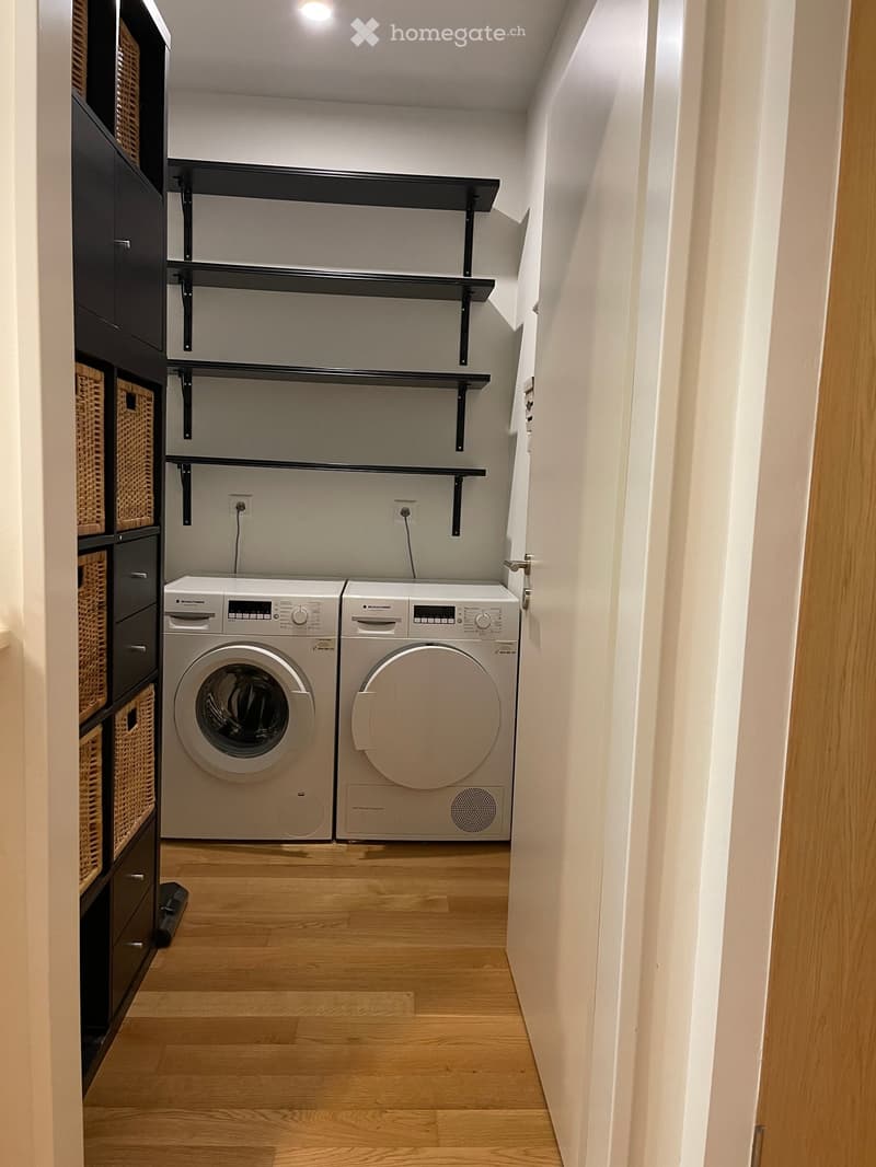 Modern laundry room with wooden flooring, white washing machine and dryer, black shelves, and wicker baskets.