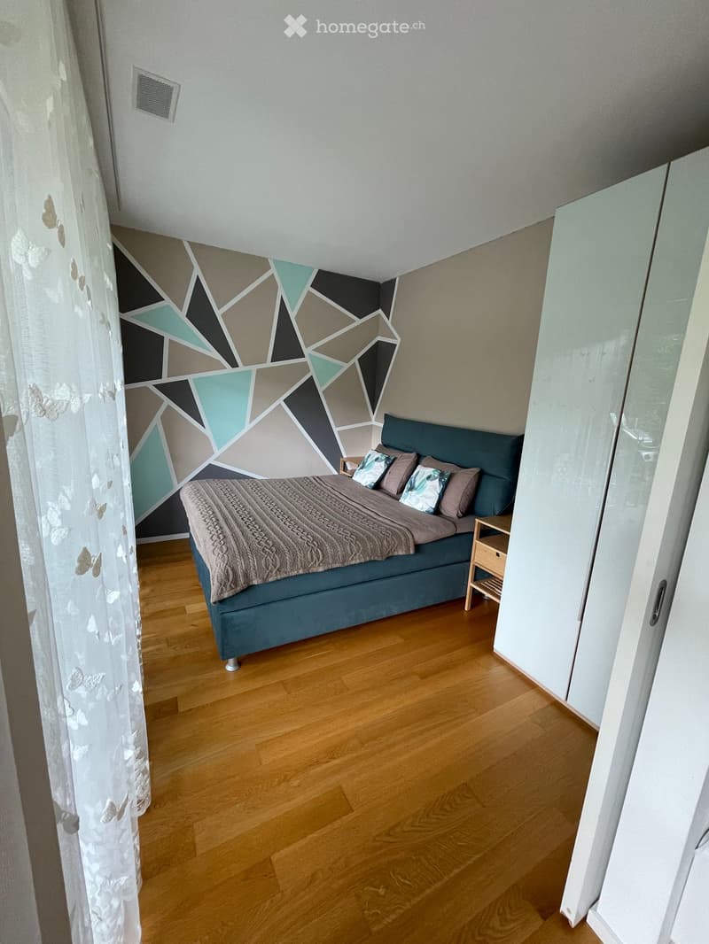Modern bedroom with wooden flooring, geometric wall design, teal headboard, and a nightstand.