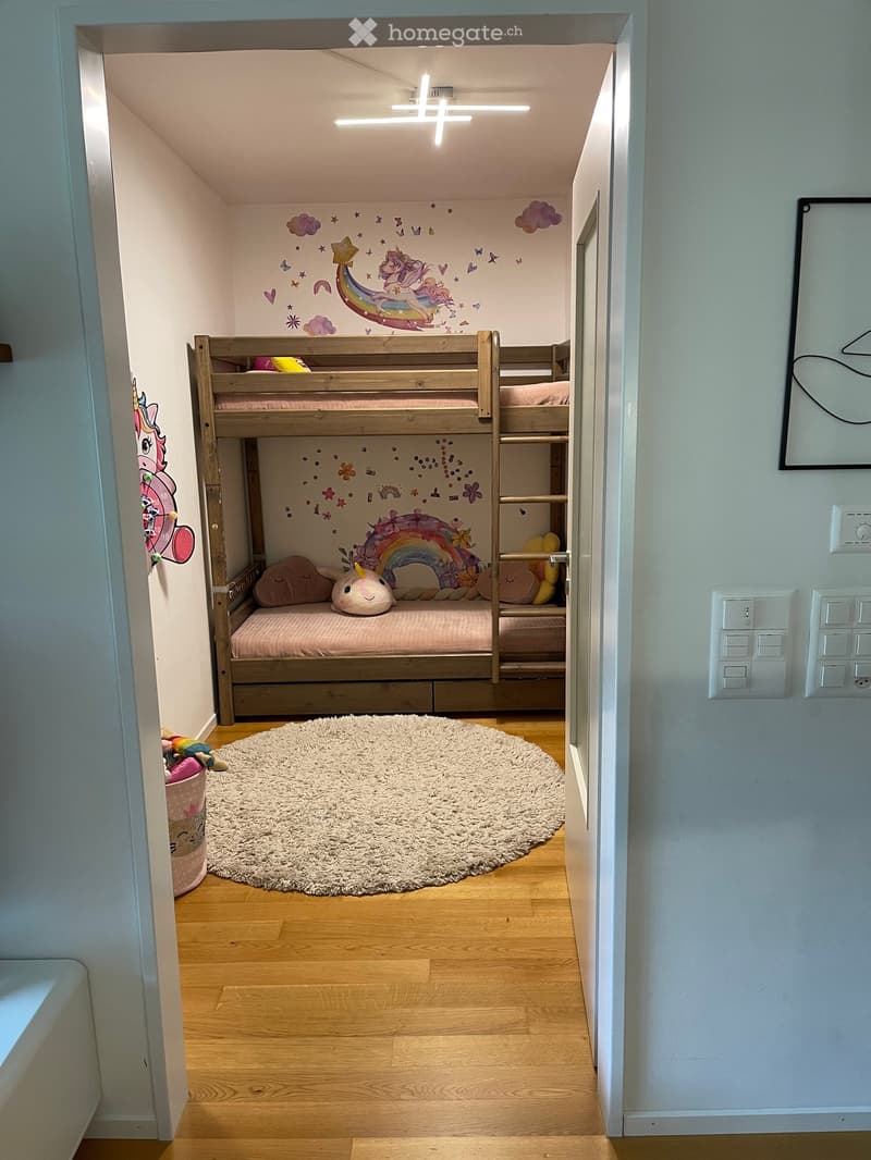 Cozy children's bedroom with a wooden bunk bed, colorful wall decals, plush toys, and a round shag rug on a wooden floor.