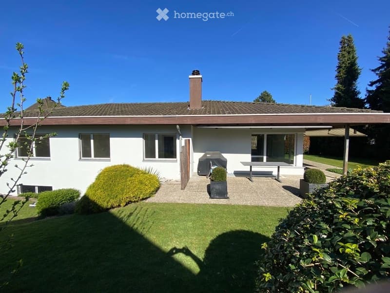 Single-story white house with a tiled roof, chimney, and large windows. The front yard features a well-maintained lawn, trimmed bushes, and a small patio area with a bench and potted plants.