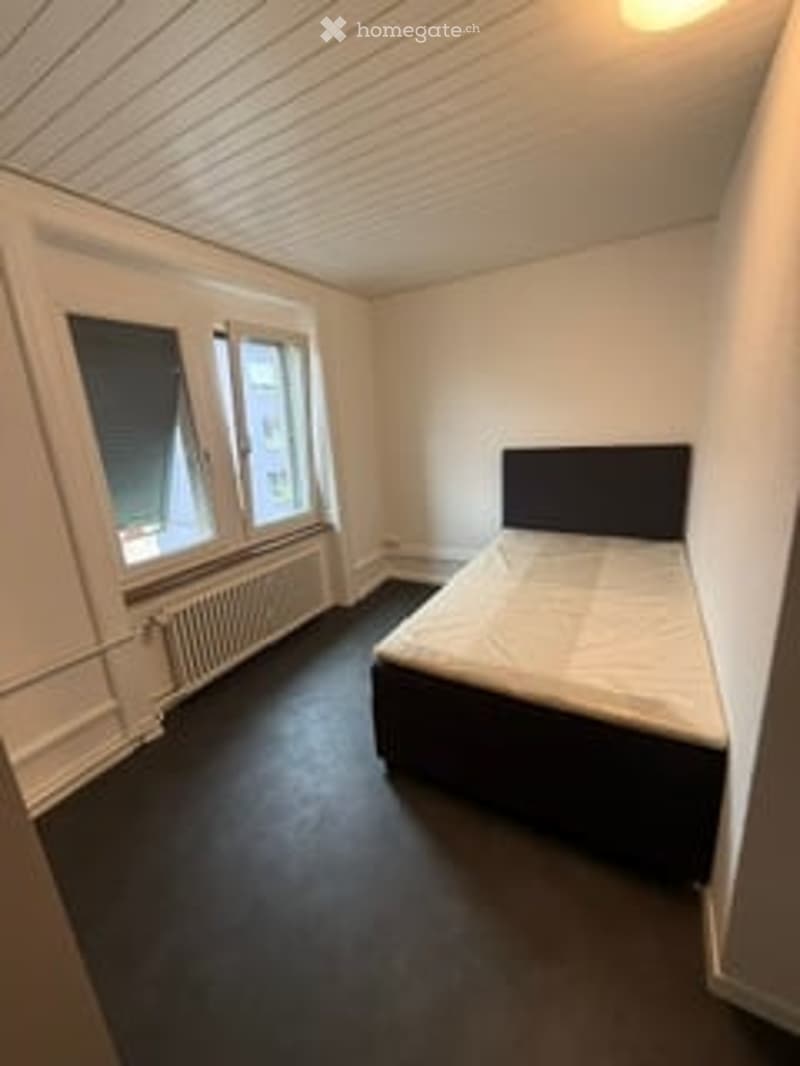 A small, unfurnished bedroom with white walls, wooden flooring, and a single bed. The room has two windows with white frames and a radiator under one of the windows.