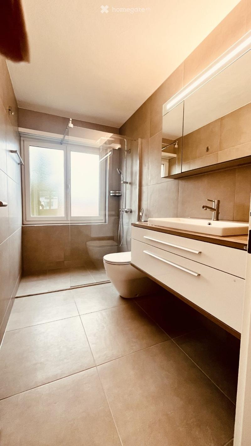 Modern bathroom with beige tiles, a large window, a toilet, a sink with a wooden vanity, and a glass-enclosed shower area.