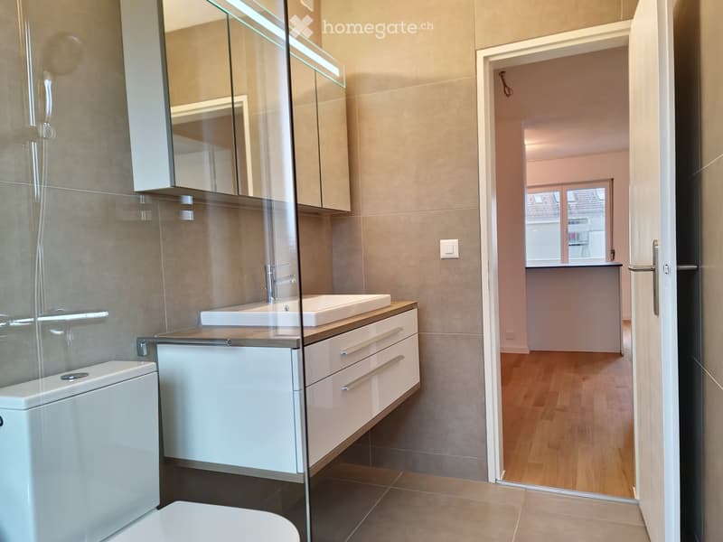 Modern bathroom with a toilet, sink, and mirrored cabinet. The room features light-colored tiles and a wooden floor visible through an open doorway.