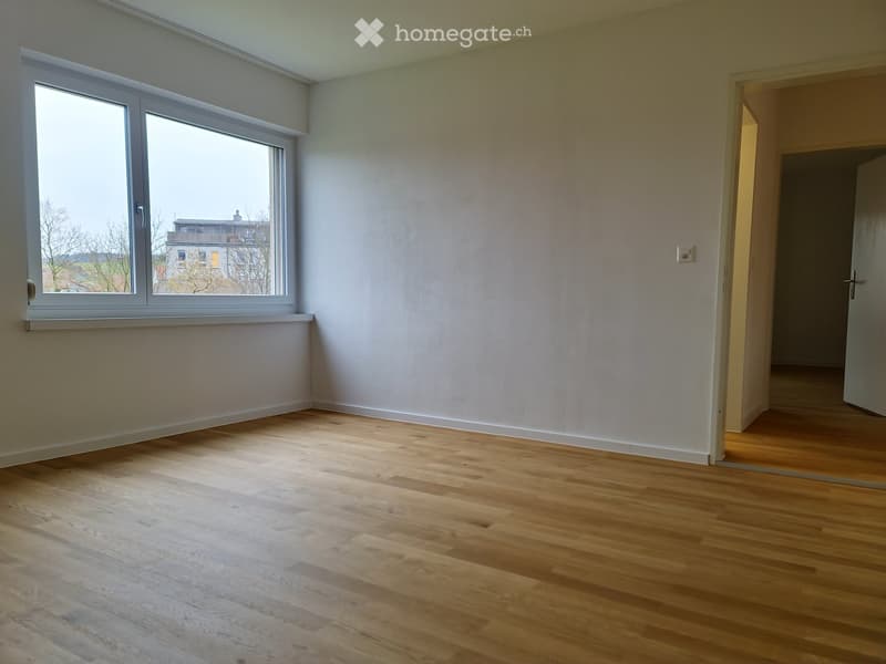 Empty room with light wooden flooring, white walls, and large windows offering a view of the outside. The room has a door leading to another space.