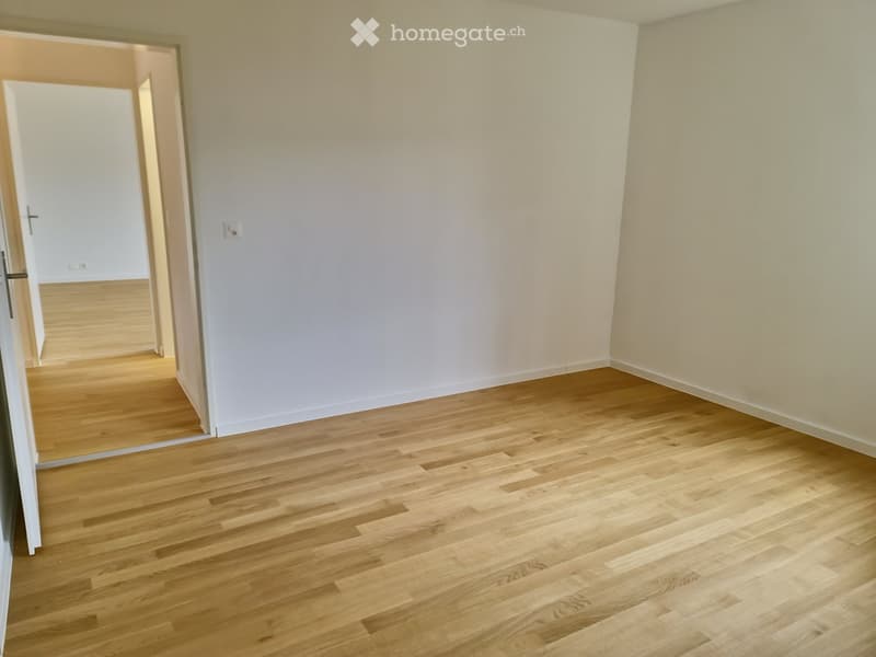 Empty room with light wooden flooring and white walls, featuring an open doorway leading to another room.