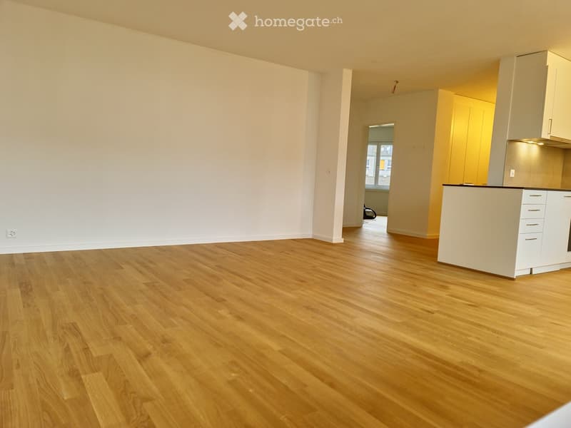 Empty room with light wooden flooring and white walls, featuring a small kitchen area with cabinets and a countertop visible in the background.