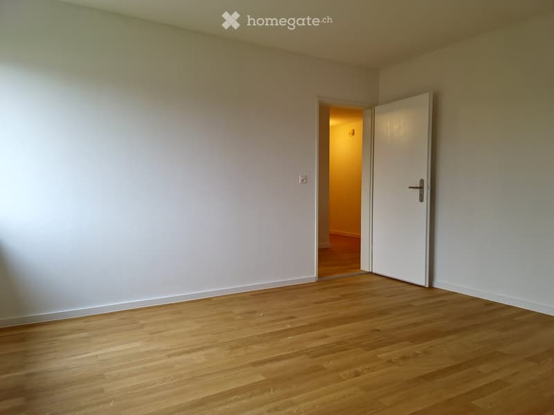 Empty room with light wooden flooring and white walls, featuring a closed white door leading to another room.