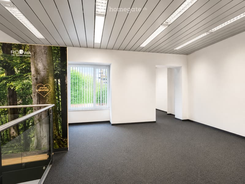Modern, empty hallway with white walls, grey carpeted floor, and a large window with blinds. A glass railing leads to an upper level with a mural of a tree and heart design.