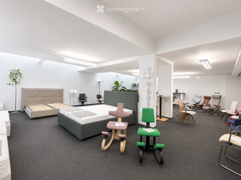 Modern showroom with two beds, various chairs, stools, and a small table, surrounded by plants and display items.