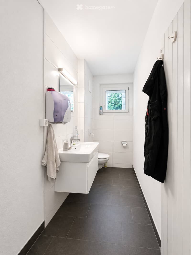 Modern bathroom with white tiles, dark grey floor, white sink, mirror, and a towel hanging on the wall. A hand dryer is mounted on the wall, and a black jacket is hanging on the right side.