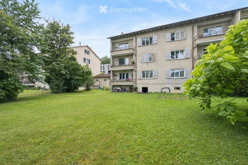 A well-maintained lawn in front of a multi-story residential building with balconies and windows. The building has a light-colored facade and is surrounded by trees and greenery.