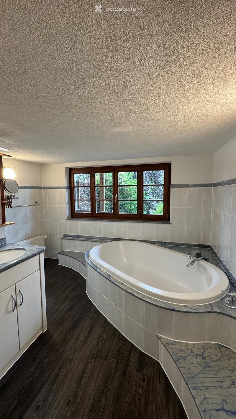 Modern bathroom with a large bathtub, sink, and mirror, featuring wooden flooring and tiled walls.