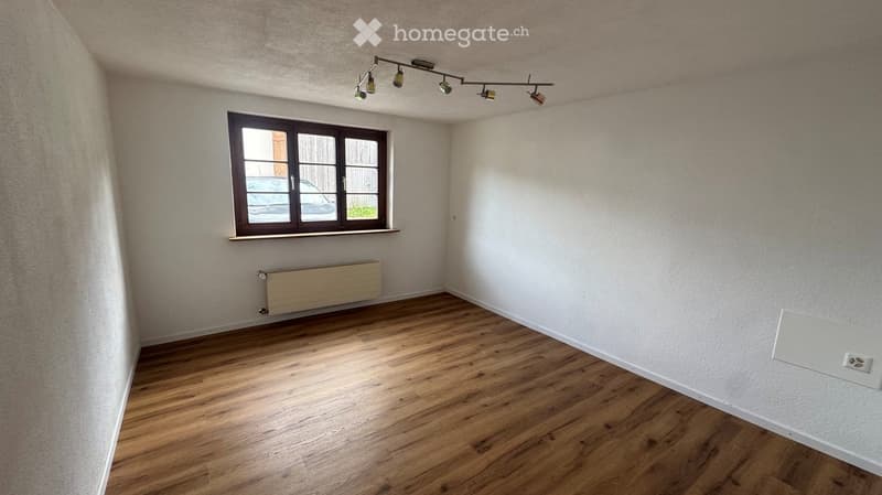 Empty room with wooden flooring, white walls, a window with a brown frame, a radiator, and a track light fixture on the ceiling.