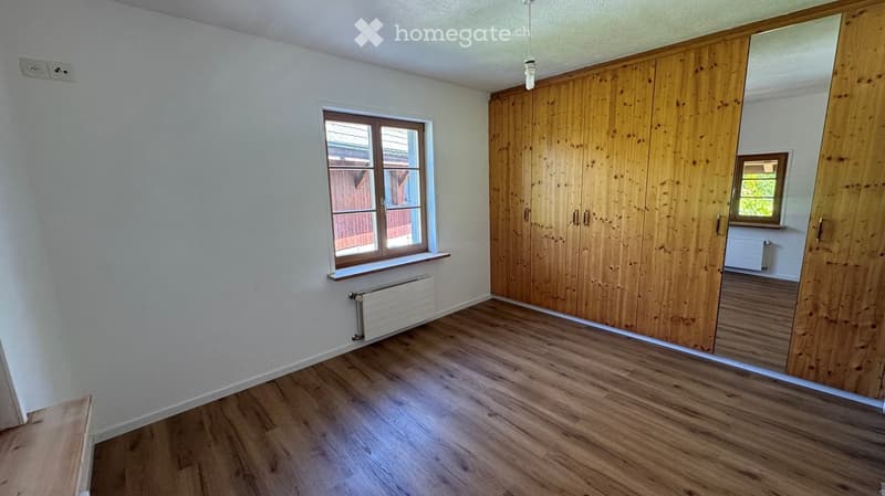 Empty room with wooden flooring, white walls, large window with wooden frame, built-in wooden closet, radiator, and electrical outlet visible.
