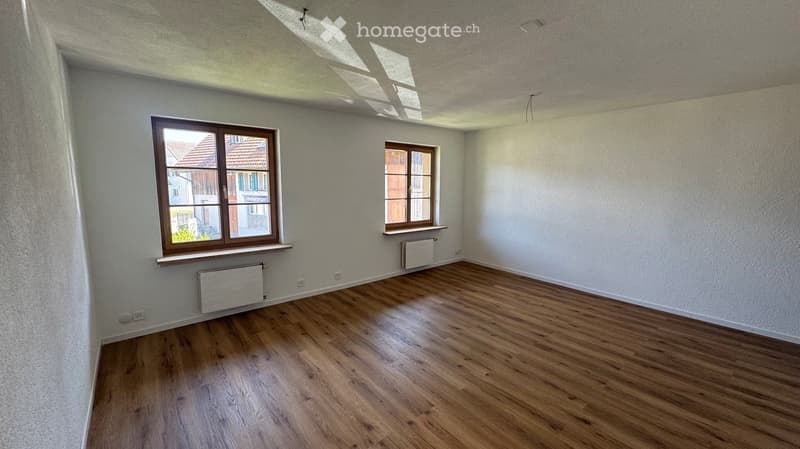 Empty room with wooden flooring, white walls, and two windows with wooden frames. Radiators are visible beneath the windows.