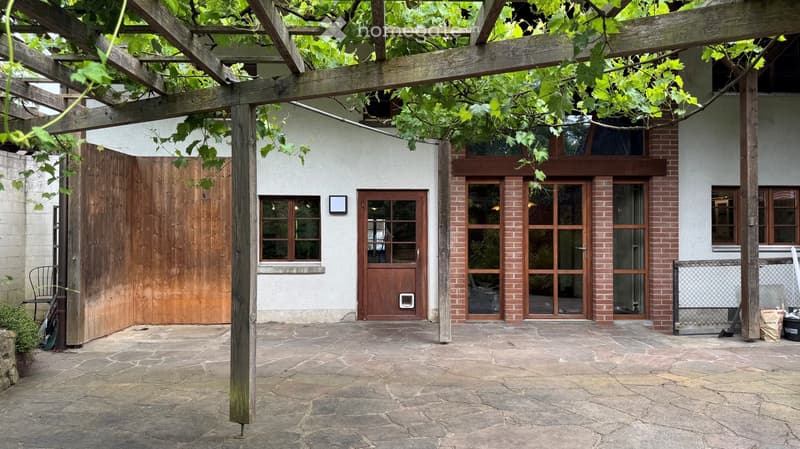 A covered outdoor area with a stone-paved ground, wooden pergola, and a building entrance with large glass doors and windows.