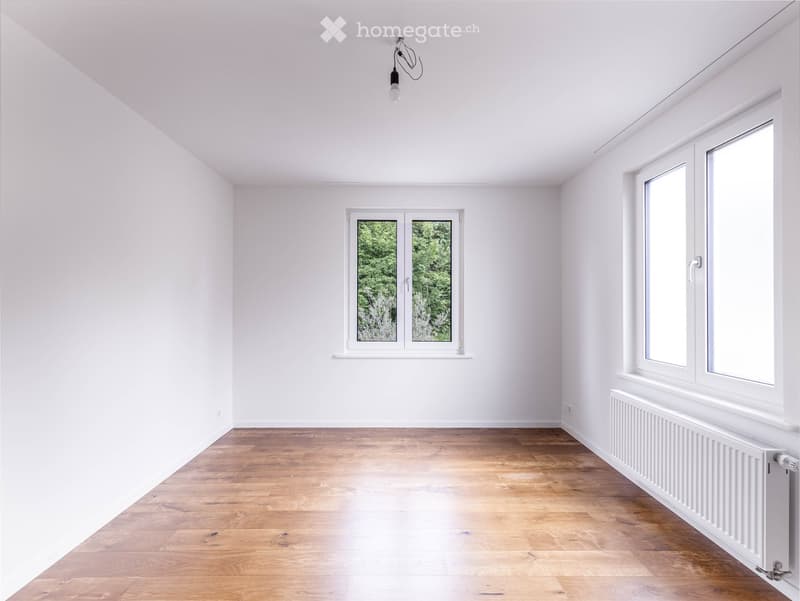 Empty room with white walls, wooden flooring, and large windows showing greenery outside. No furniture or equipment present.