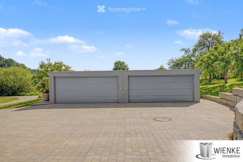Modern two-car garage with grey doors, surrounded by a paved driveway and greenery.