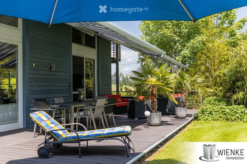 A modern outdoor patio area with a wooden deck, lounge chair, dining table with chairs, and potted plants. The patio is covered by a large retractable awning and features a view of lush greenery and a clear sky.