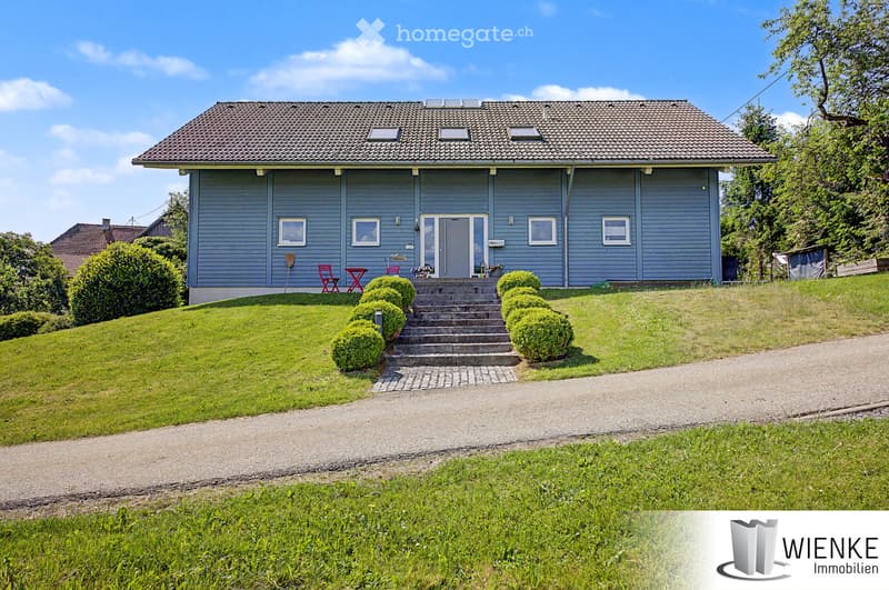 Two-story house with grey siding, multiple windows, and a front entrance with steps. The house is surrounded by a well-maintained lawn and shrubs, with a gravel driveway leading to the entrance.
