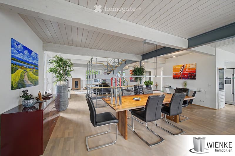 Open-plan living and dining area with wooden flooring, high exposed wooden ceiling beams, modern dining table with black leather chairs, large potted plant, artwork, and a modern kitchen area with stainless steel appliances.