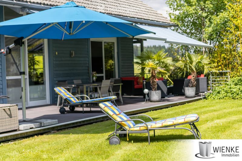 Well-maintained garden with a patio area, lounge chairs, dining table, and a large umbrella providing shade. The patio is adjacent to a house with large windows and a covered walkway.