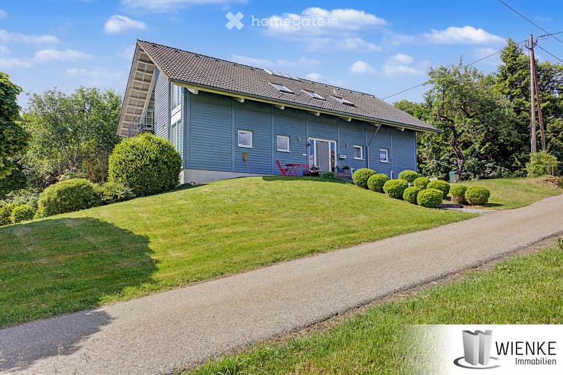 Two-story house with blue siding, multiple windows, and a raised ground floor. The house features a well-maintained lawn, trimmed bushes, and a small patio area with outdoor furniture.