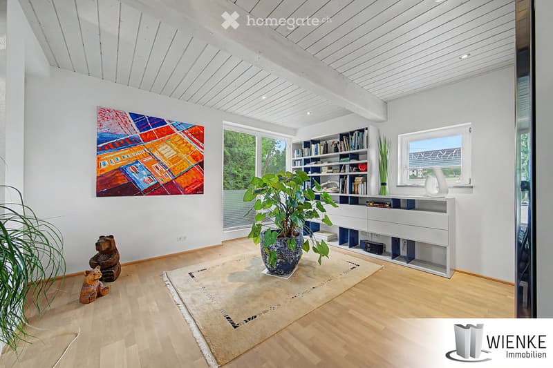 Bright living room with wooden flooring, white walls, large windows, bookshelf, potted plant, decorative vase, and artwork.