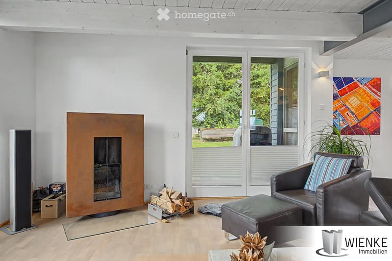 Modern living room with light wooden flooring, large windows offering a view of greenery, a black leather sofa with an ottoman, a sleek metal fireplace, a tall speaker, and decorative artwork on the wall.