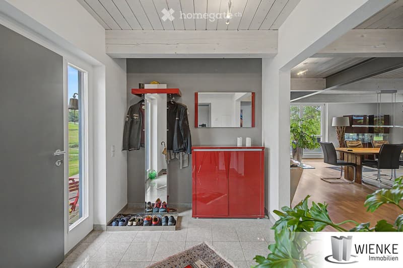 Modern hallway with white wooden ceiling beams, grey walls, red storage cabinet, coat rack with jackets, mirror, and a view into a dining area with wooden table and chairs.