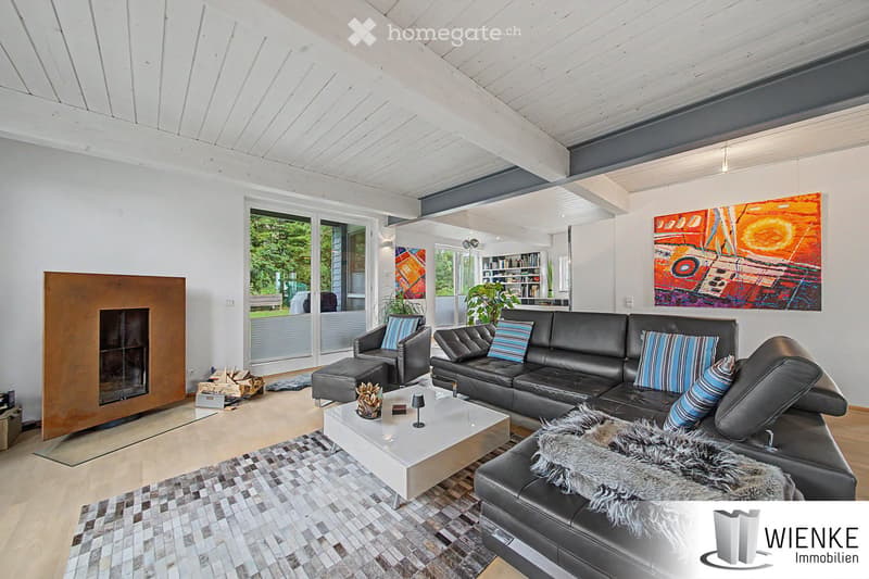 Modern living room with white walls, wooden ceiling, large windows, fireplace, sofa, coffee table, armchairs, artwork, and bookshelf.