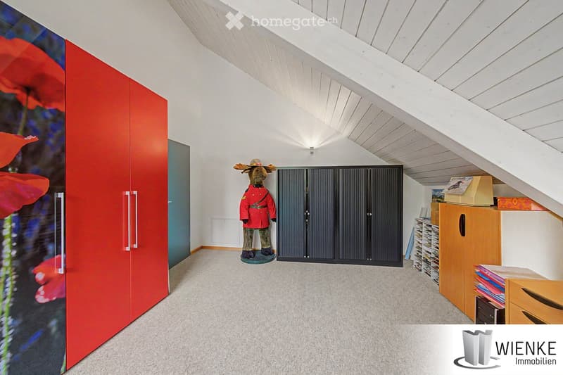 Bright, spacious room with white walls, wooden slanted ceiling, carpeted floor, large red wardrobe, grey cabinet, and storage unit with books and boxes.