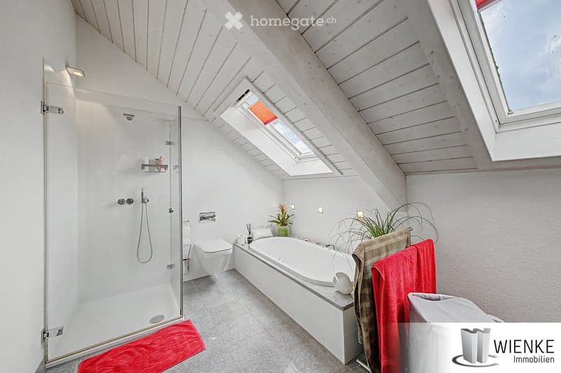 Modern bathroom with a large bathtub, separate shower, toilet, and skylight windows. Features white tiled walls, wooden ceiling beams, and decorative towels.