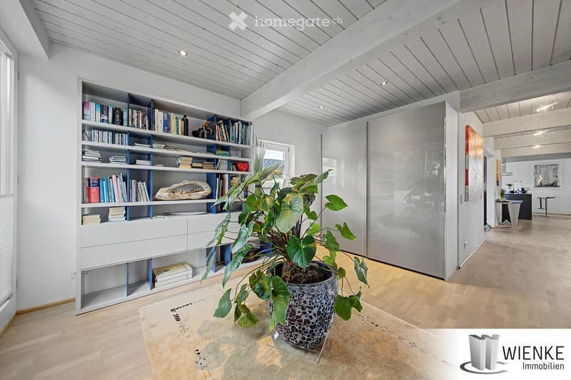 Bright, modern room with white walls, wooden floor, large bookshelf filled with books, a large leafy plant in a pot, and a small table with stools visible in the background.