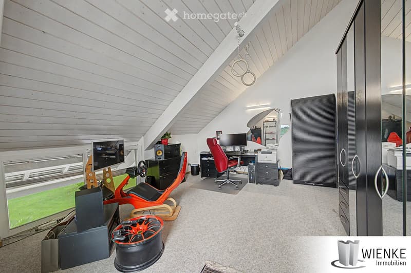 A modern, spacious room with a slanted wooden ceiling and large windows. Features include a red gaming chair, a desk with a computer monitor, a speaker system, a wheelchair, and a wheel on the floor.