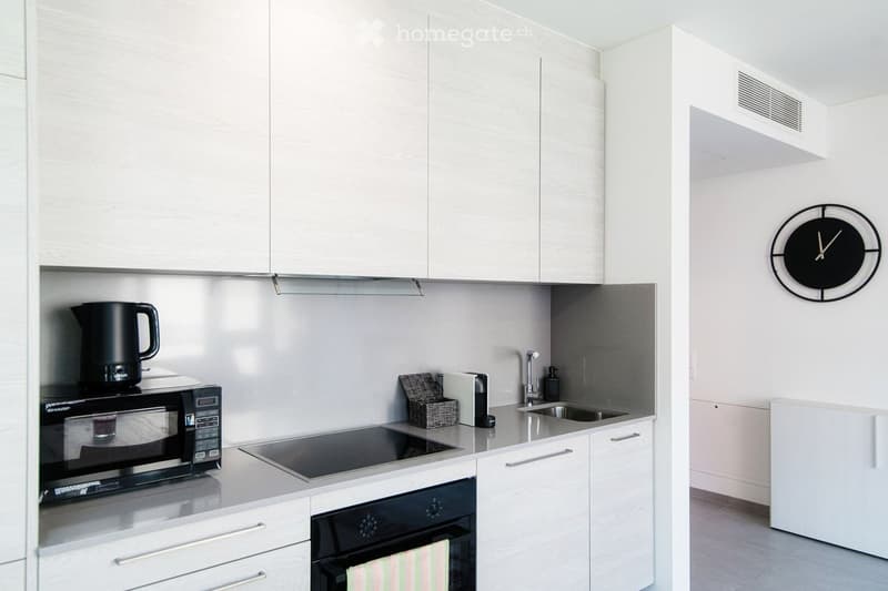 Modern kitchen with white cabinets, stainless steel appliances, and a sleek design. Features include a microwave, oven, induction stove, sink, and a wall-mounted clock.