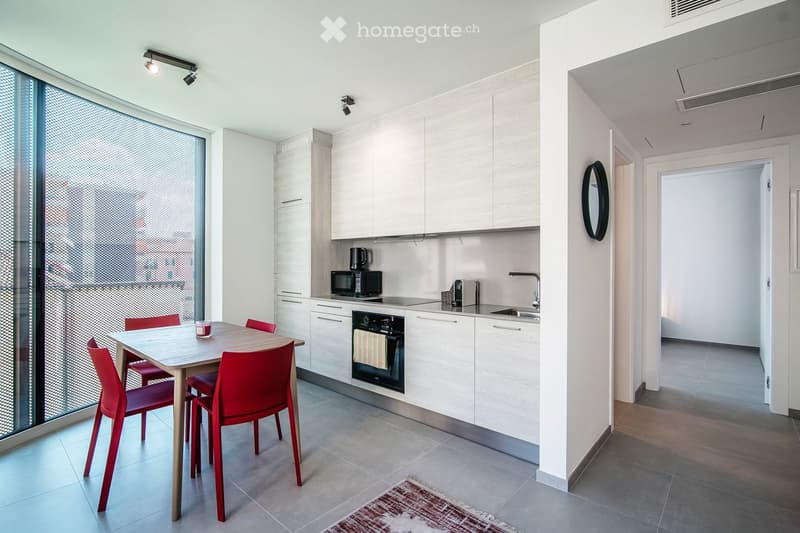 Modern kitchen with white cabinets, stainless steel appliances, a dining table with red chairs, and a large window with blinds.