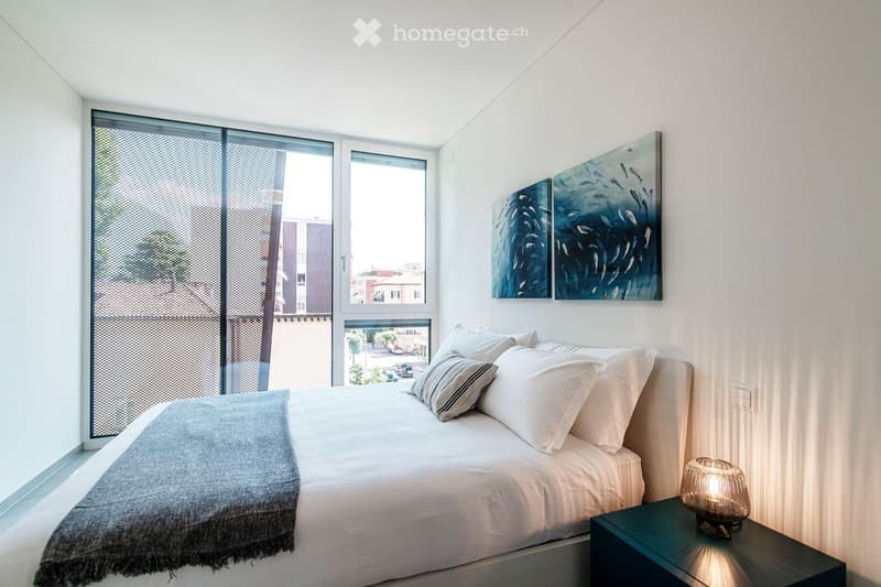Modern bedroom with a large bed, white linens, decorative pillows, a nightstand with a lamp, and a large abstract painting on the wall. Large windows with blinds offer natural light.