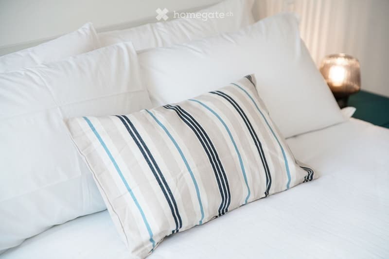 A neatly made bed with white linens and a decorative striped pillow, accompanied by a nightstand with a lamp.