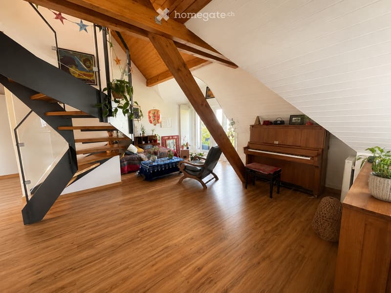 Spacious living area with wooden floors, exposed wooden beams, a grand piano, a comfortable chair, a small table, and a staircase leading to another level.