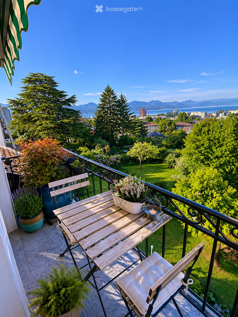 Balcony with wooden table, white cushioned chair, flower pot, and candle, overlooking a scenic view of trees, a lawn, and distant mountains.