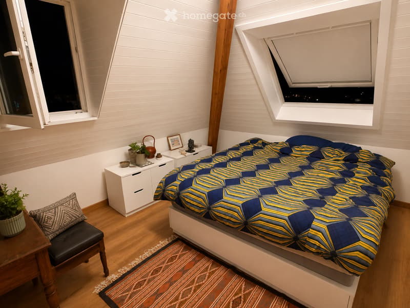 Modern bedroom with a large bed, nightstand, chair, potted plants, and decorative items. The room features wooden flooring and white paneled walls with large windows.